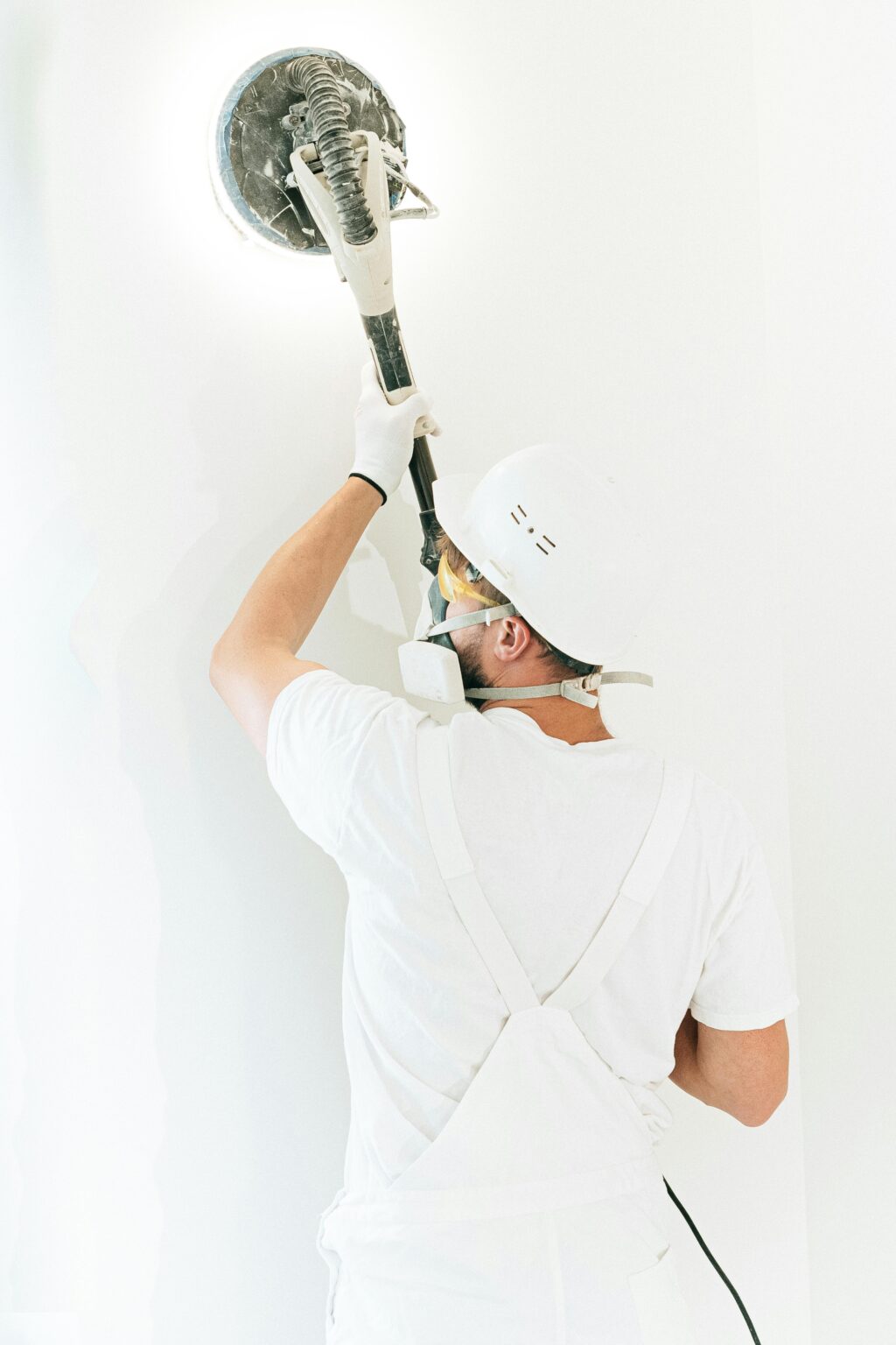 Construction worker smoothing a wall with a power sander, wearing safety gear and white overalls.